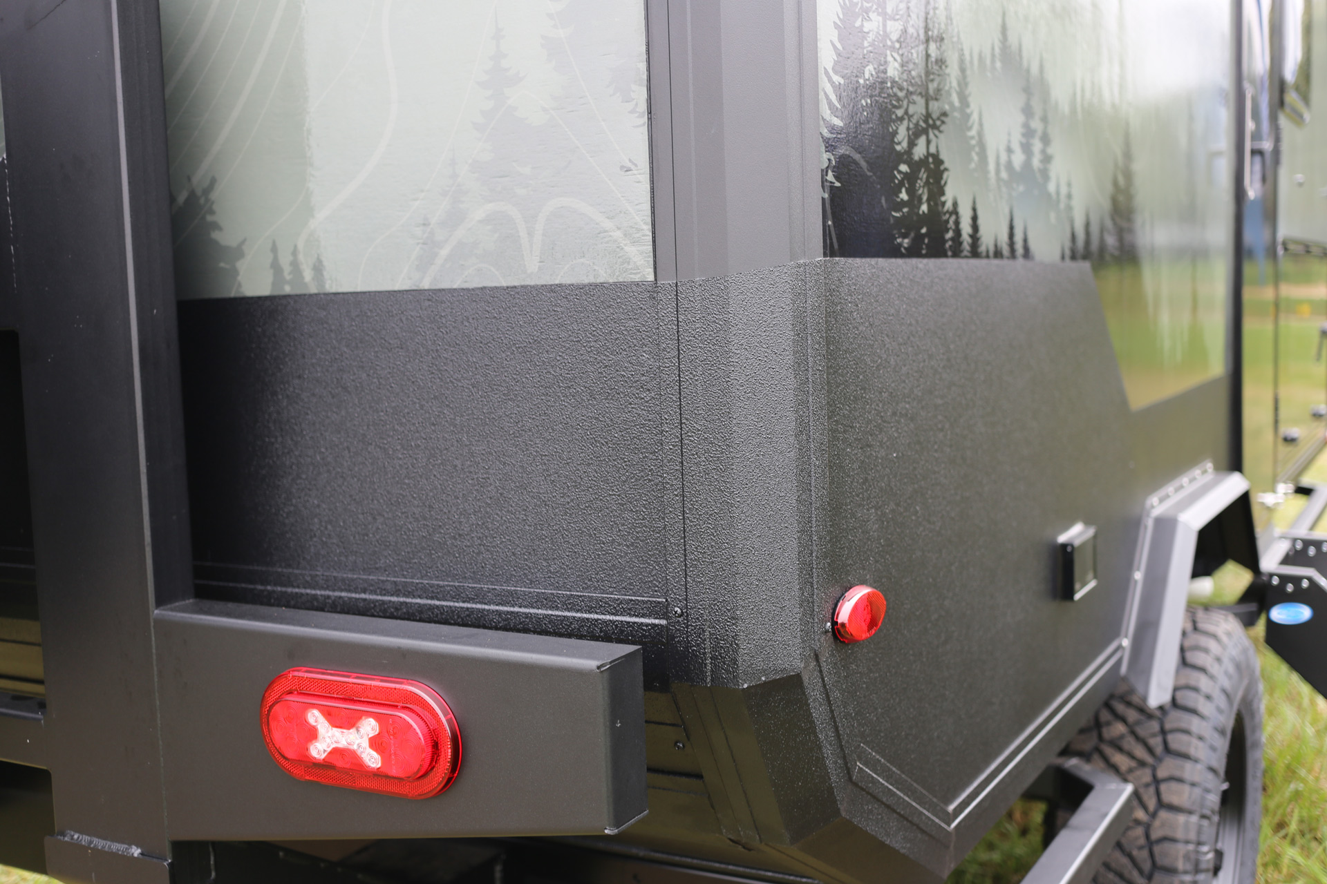 Close-up of the rear corner of a rugged off-road trailer, ideal for overland campers, featuring a textured black exterior and green side panel with a forest graphic. Visible are a small red reflector, a larger red tail light, and part of a black tire with rugged treads.