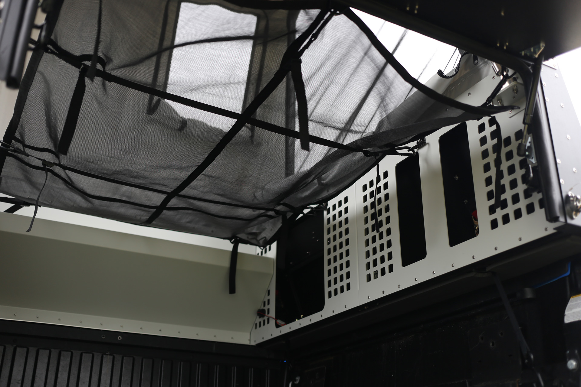 A view of the interior of an overland vehicle with a black mesh overhead storage net. The sides are lined with white and black perforated panels, showcasing the structural framework and interior storage setup ideal for Skinny Guy campers.
