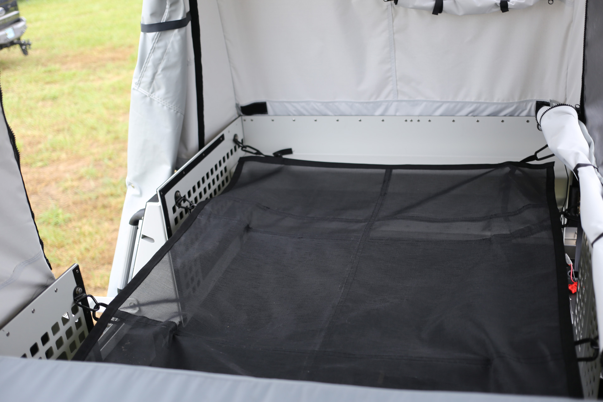 An open interior of a Skinny Guy camping trailer with a flat black mesh surface. The trailer's sides are lined with grey and black fabric panels, and the surrounding area appears to be a grassy field. This overland setup looks ready for outdoor camping or travel use.